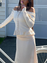 Woman wearing a polka dot off-shoulder top and beige skirt standing in front of a white door.
